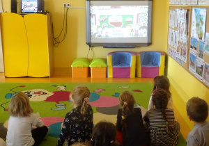 Grupa dzieci siedzi na dywanie, ogląda film edukacyjny na tablicy interaktywnej.